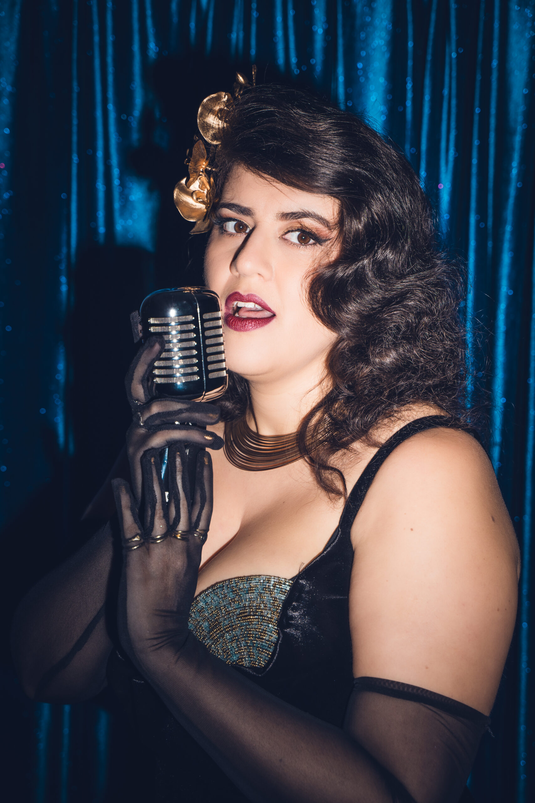 Chanteuse burlesque Cara Malice sur scène, micro vintage à la main, tenue glamour et gants noirs devant rideaux brillants.