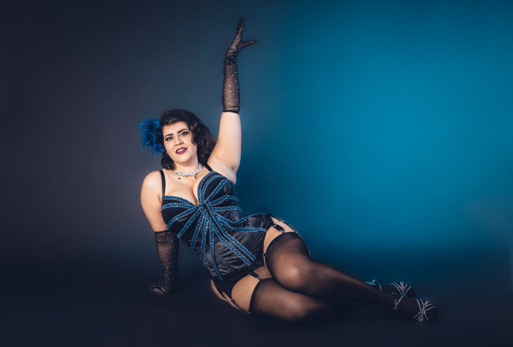 Cara Malice, artiste burlesque, pose allongée au sol en corset bleu à strass, gants longs et bas noirs, sur fond bleu studio, photographiée par Studio Of Astra.