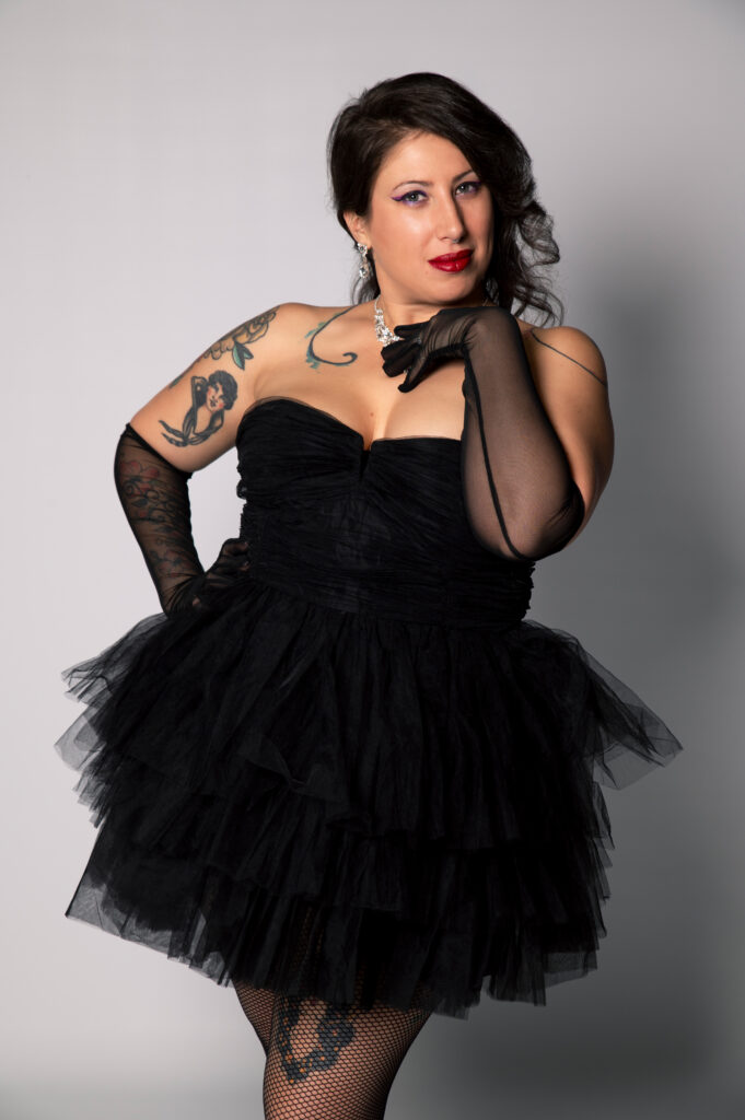 La Noushka, artiste burlesque, portant une robe bustier noire à volants en tulle, gants résille noirs, bijoux brillants et maquillage glamour, adoptant une pose élégante en studio sur fond gris, tatouages visibles.