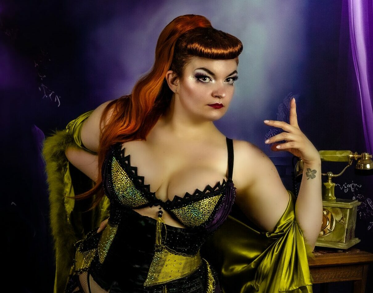 Lady Adélaïde Greed, artiste burlesque posant en corset noir, doré et violet, bas résille et kimono satiné chartreuse, devant un téléphone vintage sur une petite table dans une ambiance mystérieuse à dominante violette.