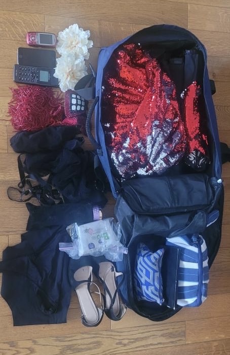 Sac ouvert contenant robe à sequins rouges, accessoires burlesques, talons, fleurs cheveux, maquillage, lingerie et objets essentiels de Cara Malice pour une performance cabaret.