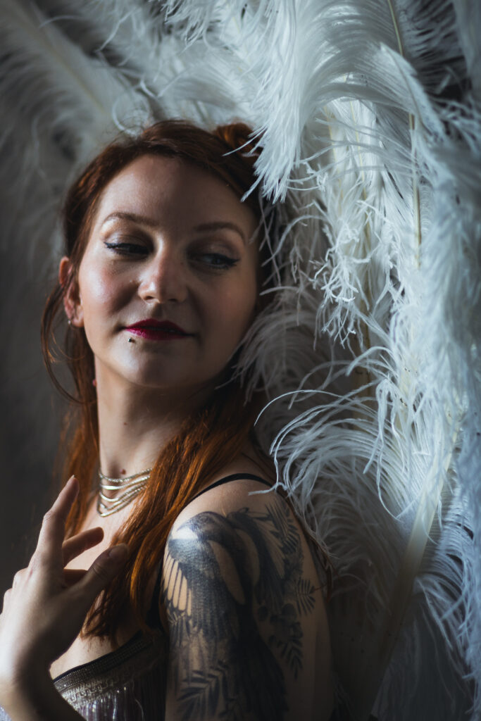 Portrait de Ness Choucrème, artiste burlesque aux cheveux roux, posant avec élégance devant un décor de plumes blanches, tatouage visible à l’épaule.