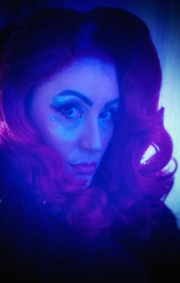 Portrait artistique d’Emeraude Plum aux cheveux ondulés violets, éclairage bleu et rose, auto-portrait burlesque.