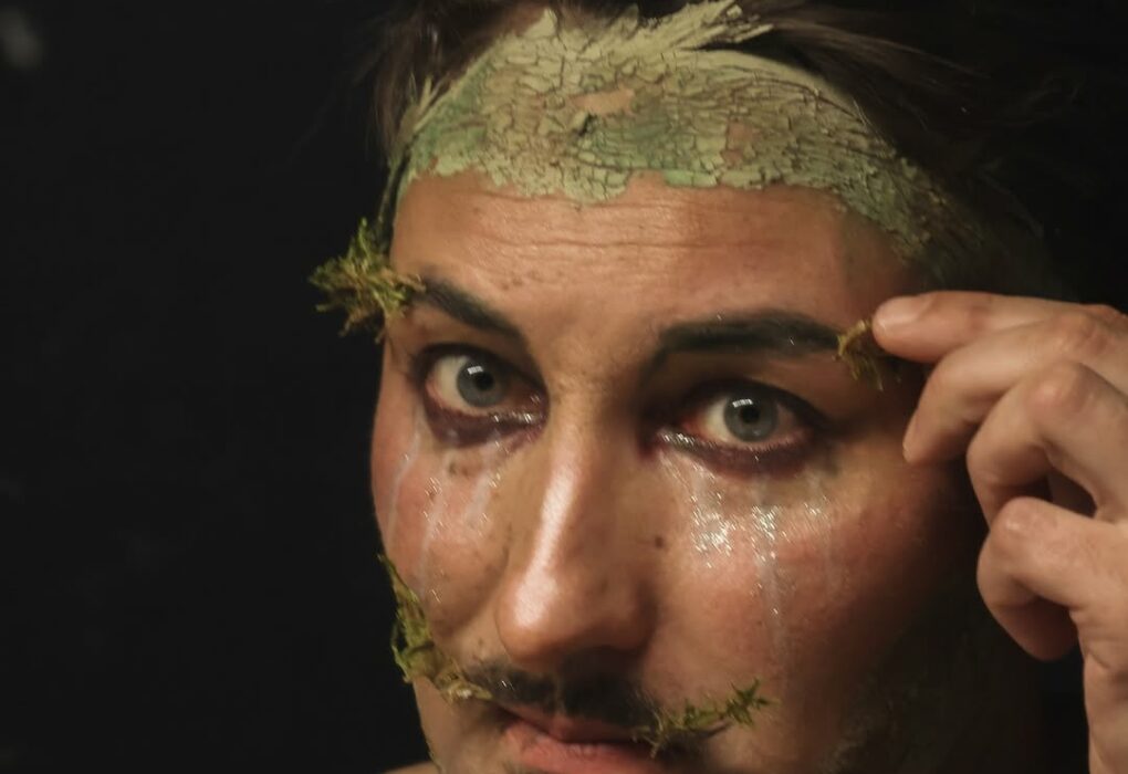 Gros plan artistique de l’artiste Gin Moustache avec le visage et le cou recouverts d’argile, sur fond sombre, sa main droite intervenant près du visage.