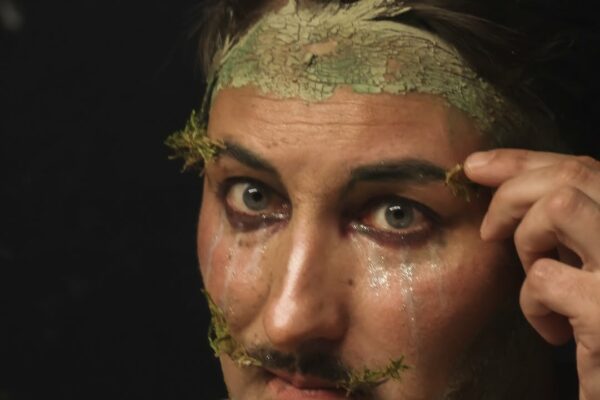 Gros plan artistique de l’artiste Gin Moustache avec le visage et le cou recouverts d’argile, sur fond sombre, sa main droite intervenant près du visage.