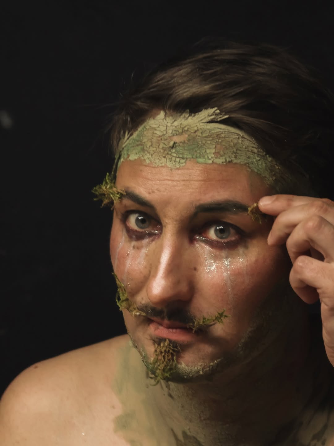 Gros plan artistique de l’artiste Gin Moustache avec le visage et le cou recouverts d’argile, sur fond sombre, sa main droite intervenant près du visage.