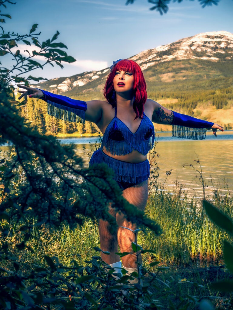 Chérie Coquette dans un costume burlesque bleu à franges, posant en pleine nature devant un lac et des montagnes, cliché signé Kathryn Nickford
