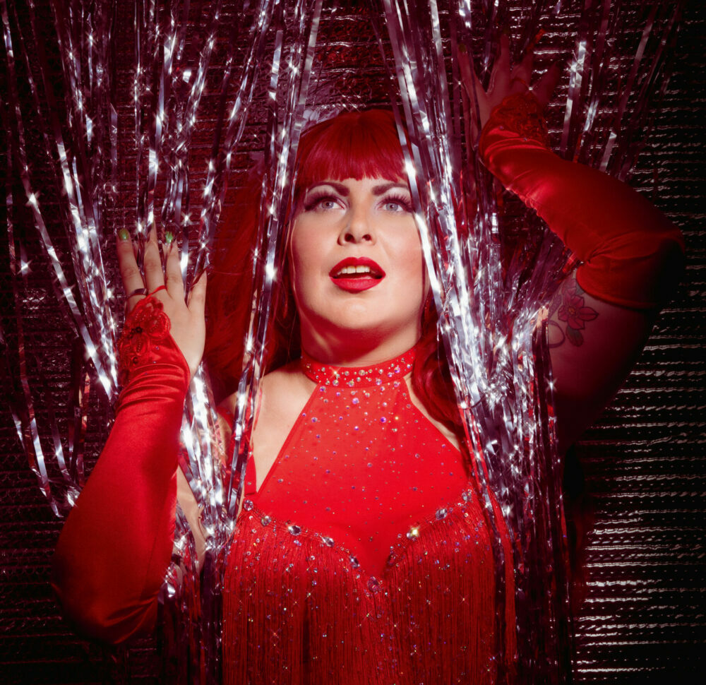 Artiste burlesque Chérie Coquette dans une tenue rouge à franges, posant devant une décoration scintillante lors d’un shooting réalisé par Jonathan-Serge Lalande.