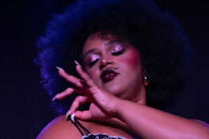 Gros plan de Vénus de Mai sur scène, portant un ensemble de lingerie satinée claire avec volants contrastés, la main levée devant le visage, éclairée par une lumière violette.