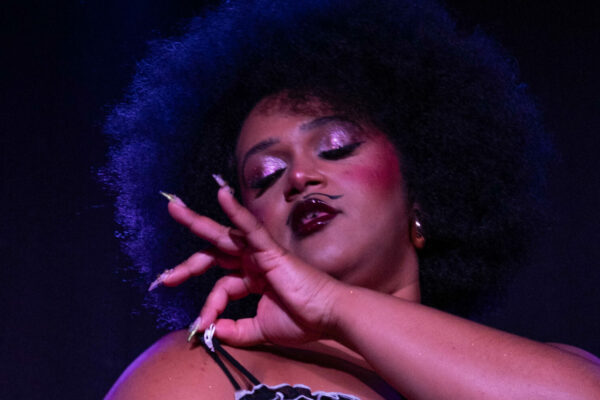 Gros plan de Vénus de Mai sur scène, portant un ensemble de lingerie satinée claire avec volants contrastés, la main levée devant le visage, éclairée par une lumière violette.