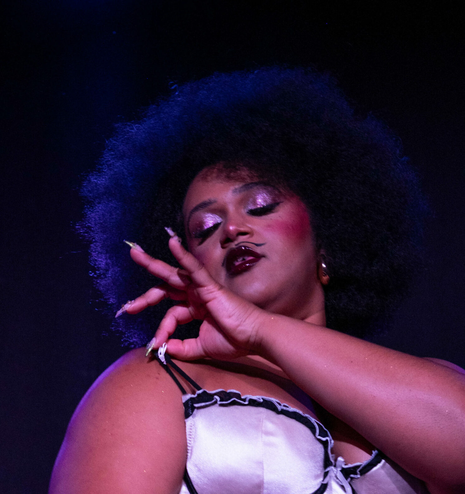 Gros plan de Vénus de Mai sur scène, portant un ensemble de lingerie satinée claire avec volants contrastés, la main levée devant le visage, éclairée par une lumière violette.