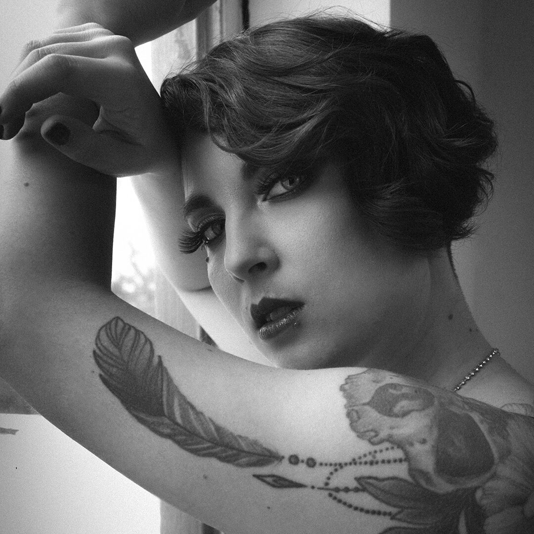 Portrait en noir et blanc de Didi Pattes de Velours, artiste burlesque, au regard intense, avec tatouage plume sur le bras. Photo Eve Saint-Ramon.
