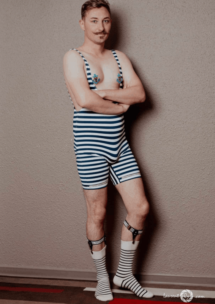 Artiste boylesque Gin Moustache posant debout en maillot de bain rétro rayé bleu et blanc, avec bretelles, jarretelles et chaussettes coordonnées, photographié en pied contre un mur neutre.