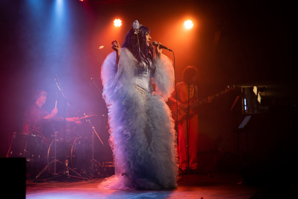 Emeraude Plum en costume nuageux sur scène, chante avec musiciens sous lumière colorée, photo de Mathieu Genon.