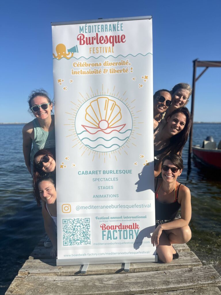 Équipe du Méditerranée Burlesque Festival posant au bord de l’eau avec la bannière officielle de l’événement, organisé par Boardwalk Factory.