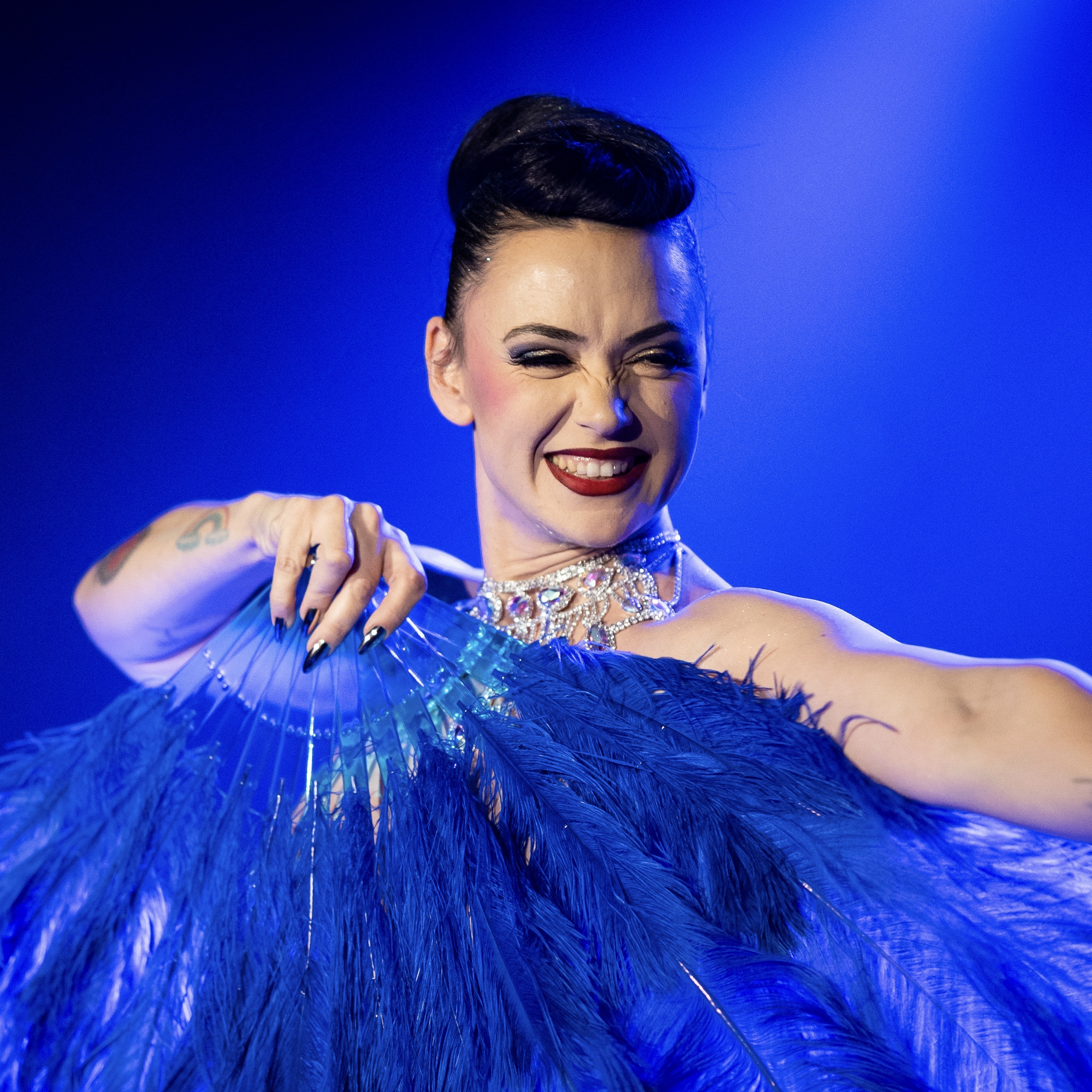 Performeur burlesque MILF Ta Mère tenant un grand éventail bleu sur scène, portrait par David Mançois.