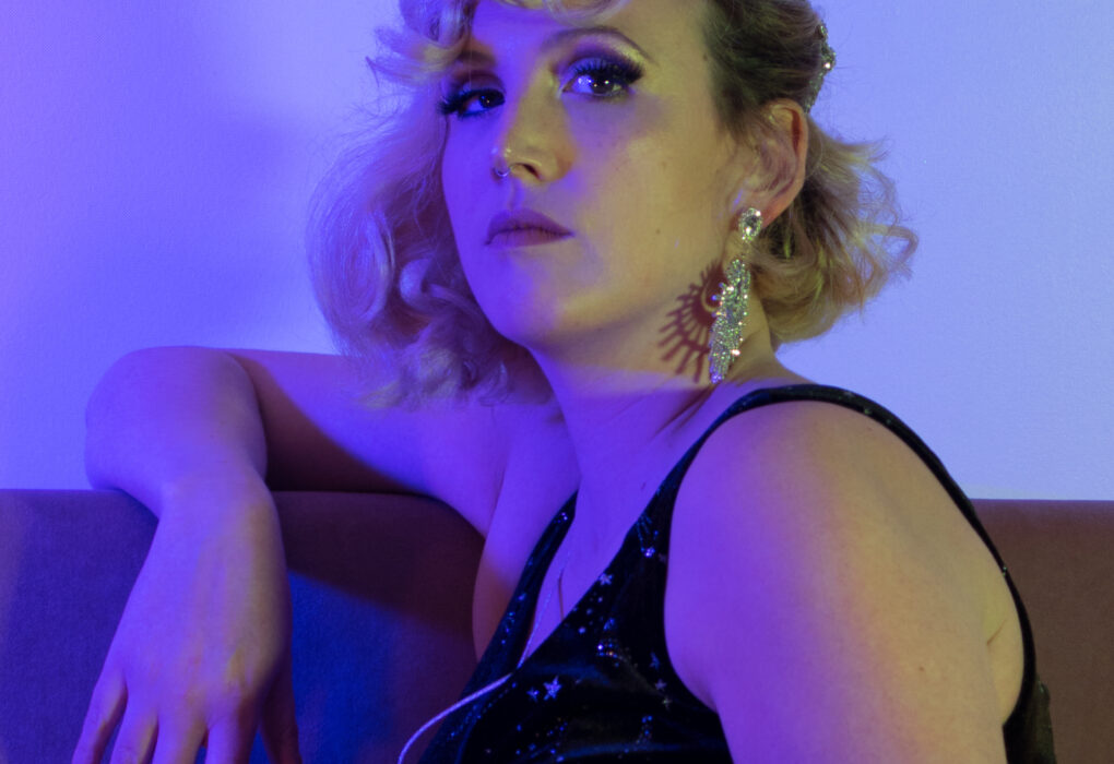 Artiste burlesque Léo Poldine posant en robe noire pailletée sur un sofa, lumières violettes – portrait par Alexia Zamuner.