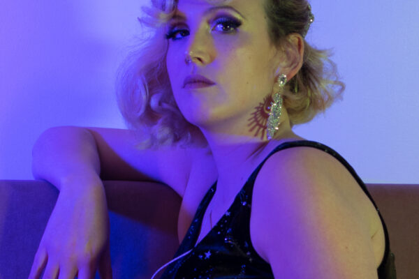 Artiste burlesque Léo Poldine posant en robe noire pailletée sur un sofa, lumières violettes – portrait par Alexia Zamuner.