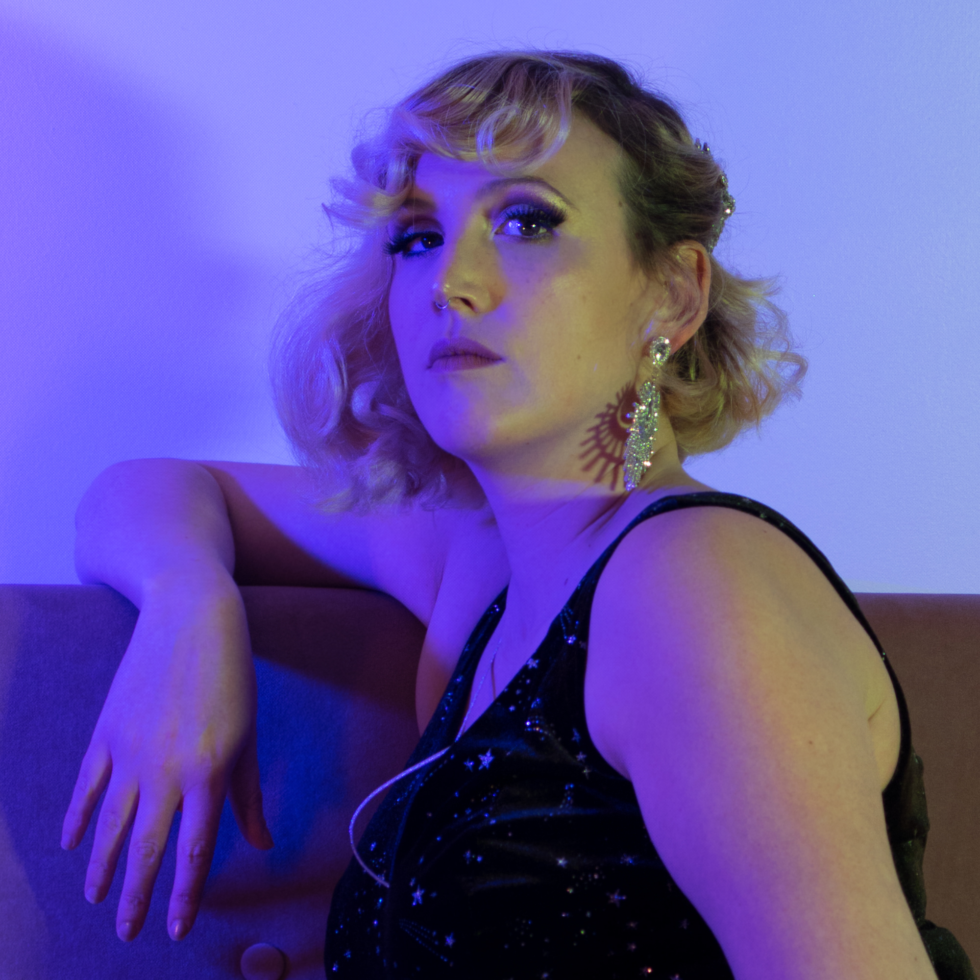 Artiste burlesque Léo Poldine posant en robe noire pailletée sur un sofa, lumières violettes – portrait par Alexia Zamuner.