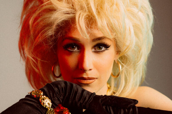 Gros plan de l’artiste burlesque Lolita Laze avec une coiffure blonde volumineuse, portant des gants noirs et des bijoux dorés, appuyée sur une surface réfléchissante.