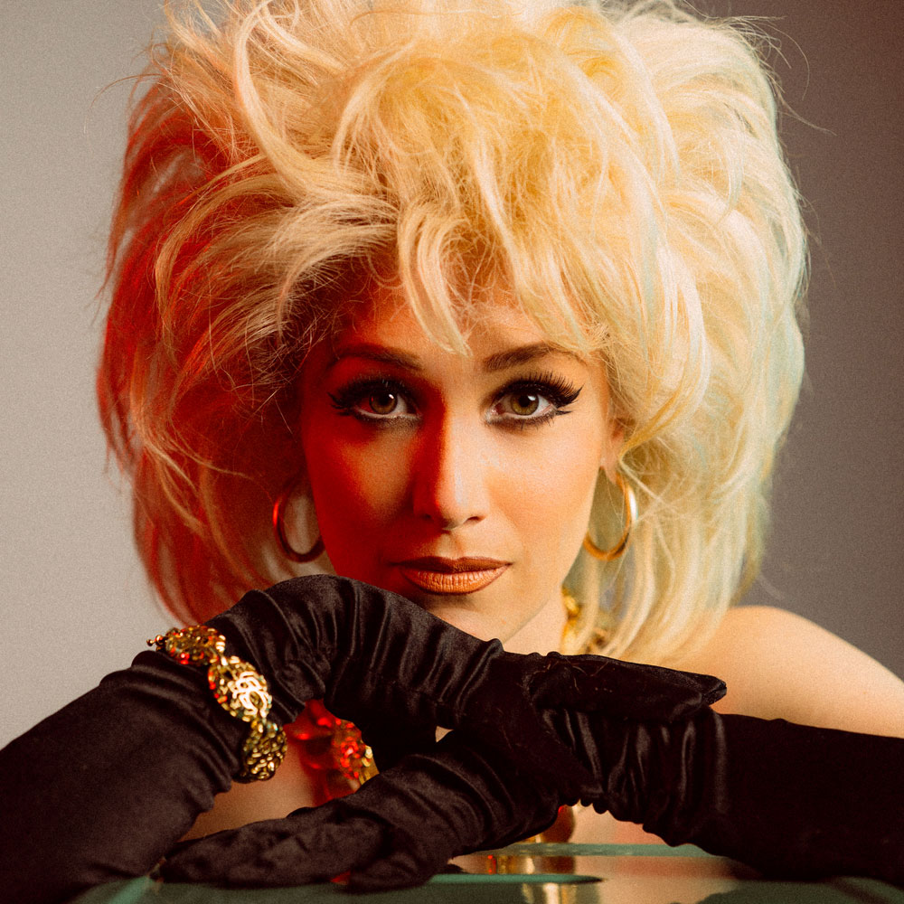 Gros plan de l’artiste burlesque Lolita Laze avec une coiffure blonde volumineuse, portant des gants noirs et des bijoux dorés, appuyée sur une surface réfléchissante.