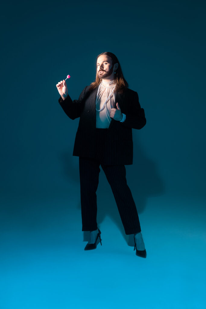 Portrait artistique de Mattamore en costume sombre, portant des perles et tenant une sucette rose, éclairée par une lumière bleutée. »