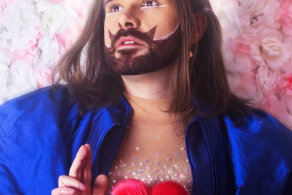 Portrait artistique d’une personne allongée sur un fond floral rose, vêtue d’un tissu bleu vif et portant un cœur rouge sur le torse.