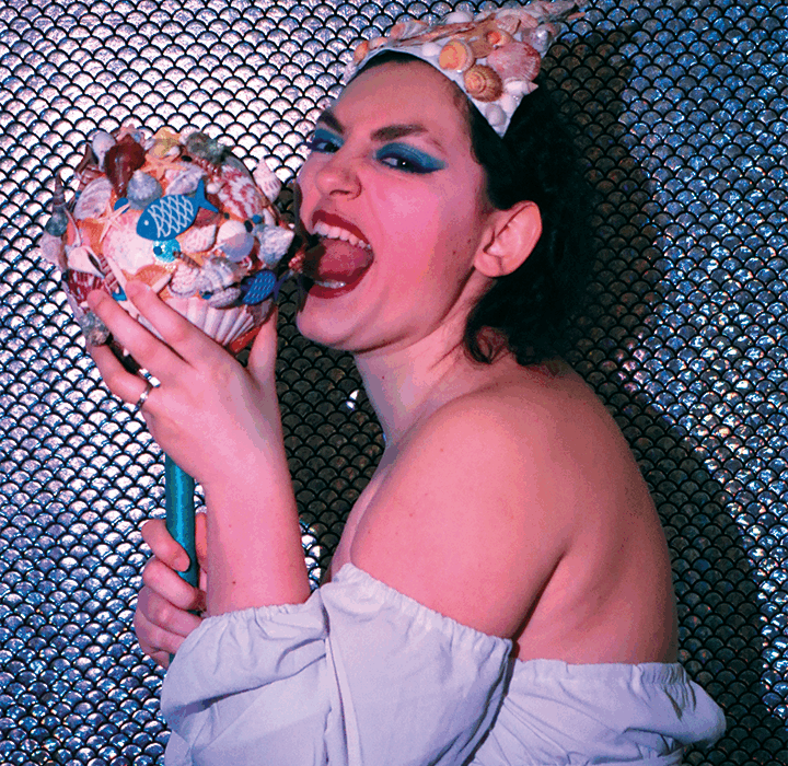 Pink Amanite avec une coiffe de coquillages, maquillage bleu et top blanc, mordant un sceptre décoré de coquillages sur fond scintillant