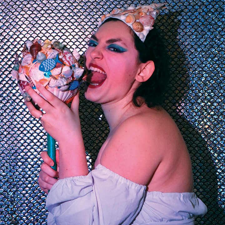 Pink Amanite avec une coiffe de coquillages, maquillage bleu et top blanc, mordant un sceptre décoré de coquillages sur fond scintillant