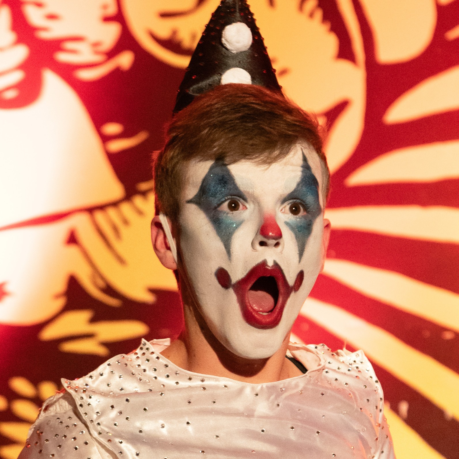 Portrait scénique de l’artiste burlesque L’innommable, vêtu d’un costume de clown contemporain serti de strass et coiffé d’un chapeau pointu, lors d’une performance haute en couleur sur scène. Crédit photo : Loraosa Pin-up.