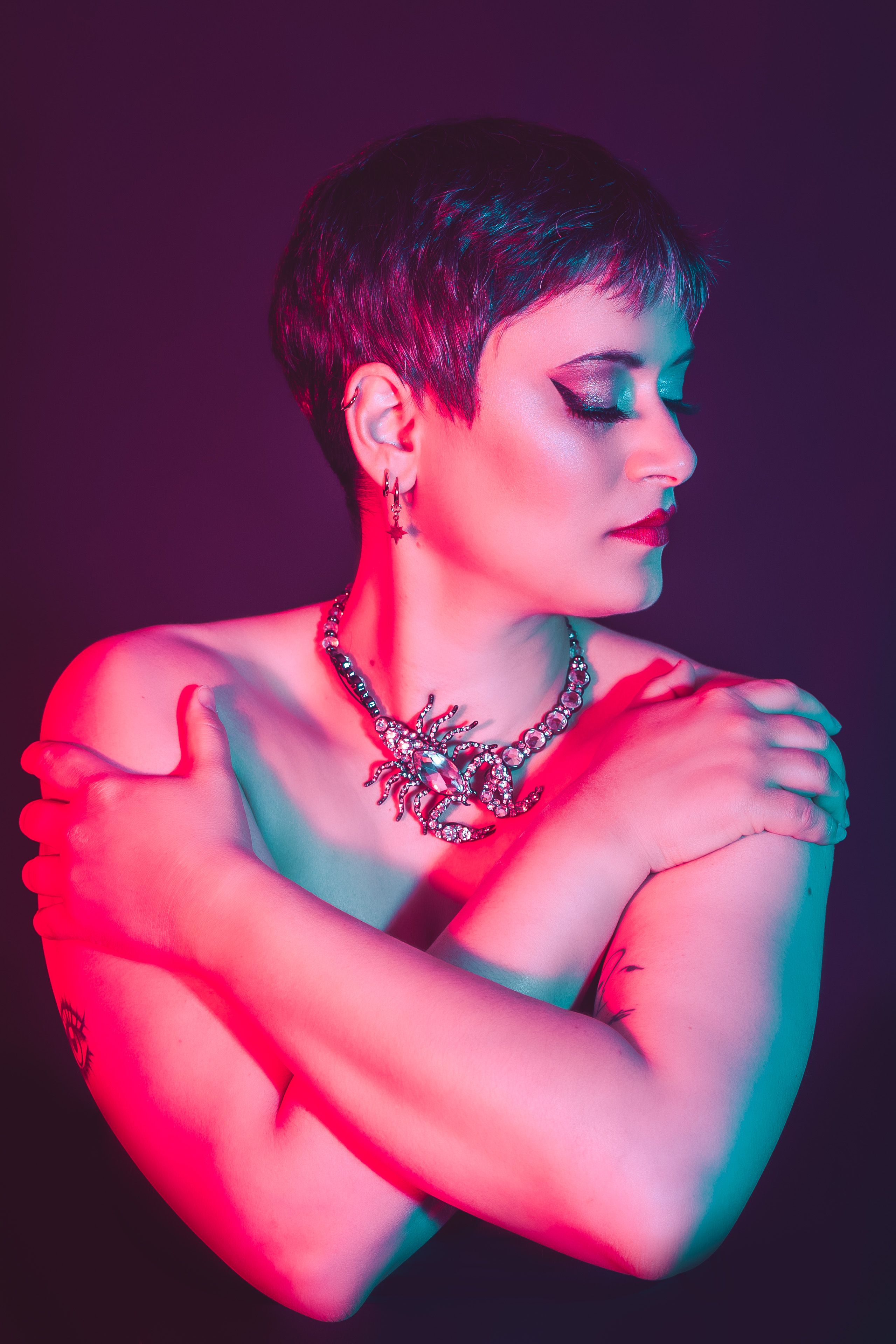 Personne aux cheveux courts, éclairée de lumières rose et bleu, portant un collier scorpion, pose artistique photographiée par Studio Of Astra.
