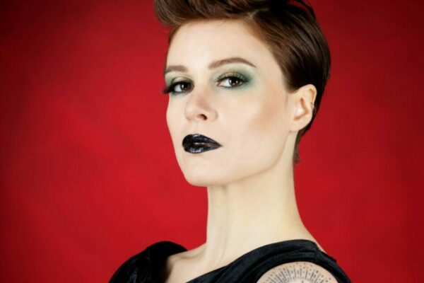 Portrait de Zazou Minaude, artiste burlesque, de profil avec tatouages sur bras et robe noire sur fond rouge, photo réalisée par Eden Concept Studio.