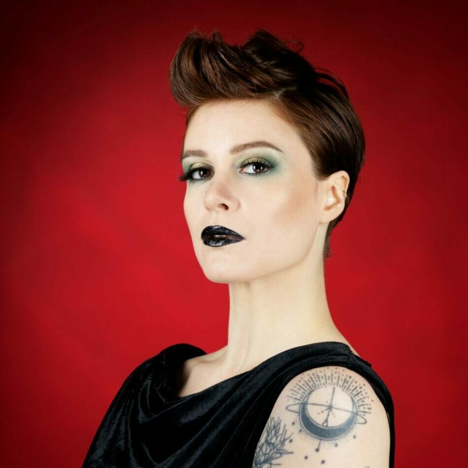 Portrait de Zazou Minaude, artiste burlesque, de profil avec tatouages sur bras et robe noire sur fond rouge, photo réalisée par Eden Concept Studio.