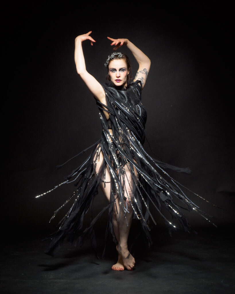 Zazou Minaude artiste burlesque, en pleine pose dans une tenue noire à franges sur fond noir, photographie signée Patrick.photoss.