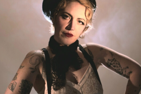 Portrait scénique d’Anita Gein, artiste burlesque, éclairée à contre‑jour, portant débardeur en dentelle, bretelles noires, béret et tatouages visibles sur les bras.