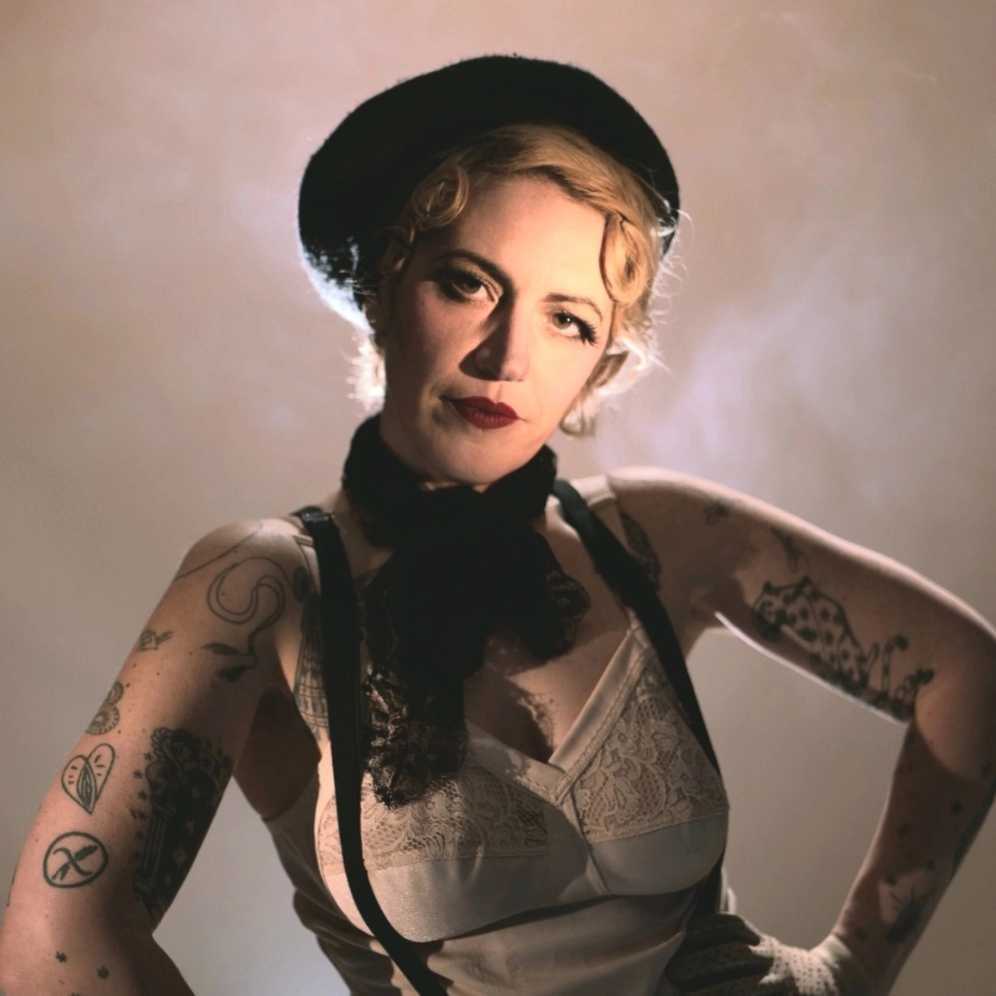 Portrait scénique d’Anita Gein, artiste burlesque, éclairée à contre‑jour, portant débardeur en dentelle, bretelles noires, béret et tatouages visibles sur les bras.