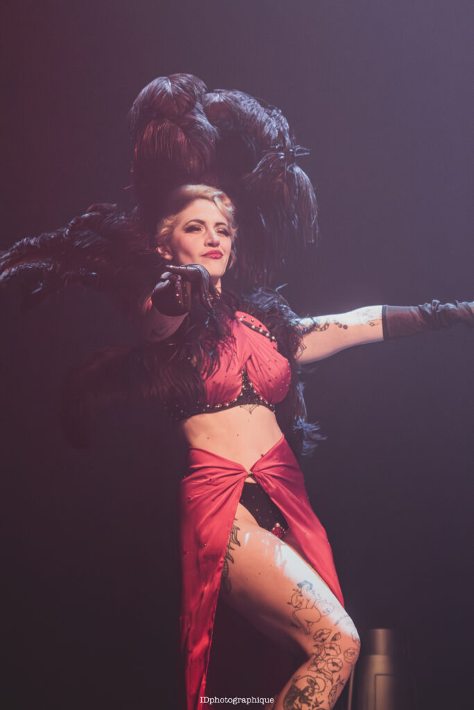 Portrait d’Anita Gein, artiste burlesque, en gros plan scénique, portant débardeur clair et bretelles noires, éclairée par une lumière de spectacle.