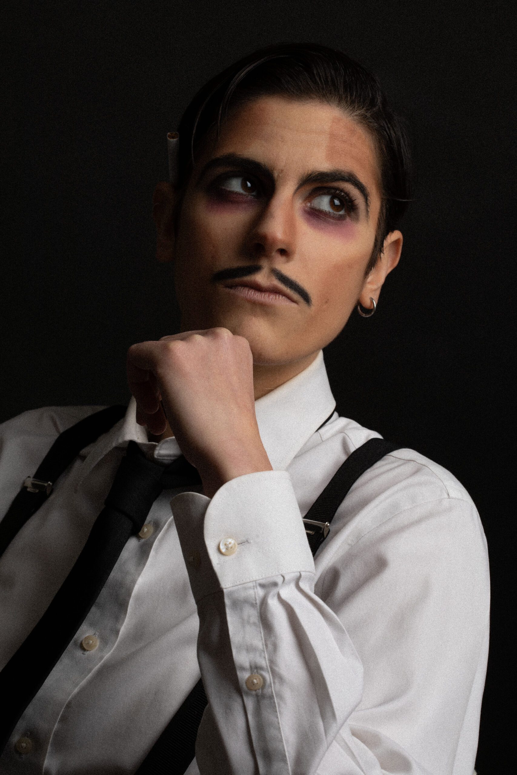 Portrait de Rico Tourky-Brille en tenue de drag king, chemise blanche, cravate noire et bretelles, posant de profil sur fond sombre, photographie de Pauline Lionne Bessaudou.