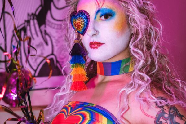 Saccage Dorée en tenue burlesque arc-en-ciel, maquillage coloré et cache-œil en cœur pailleté, posant dans une lumière rose vive – Photo Léo White