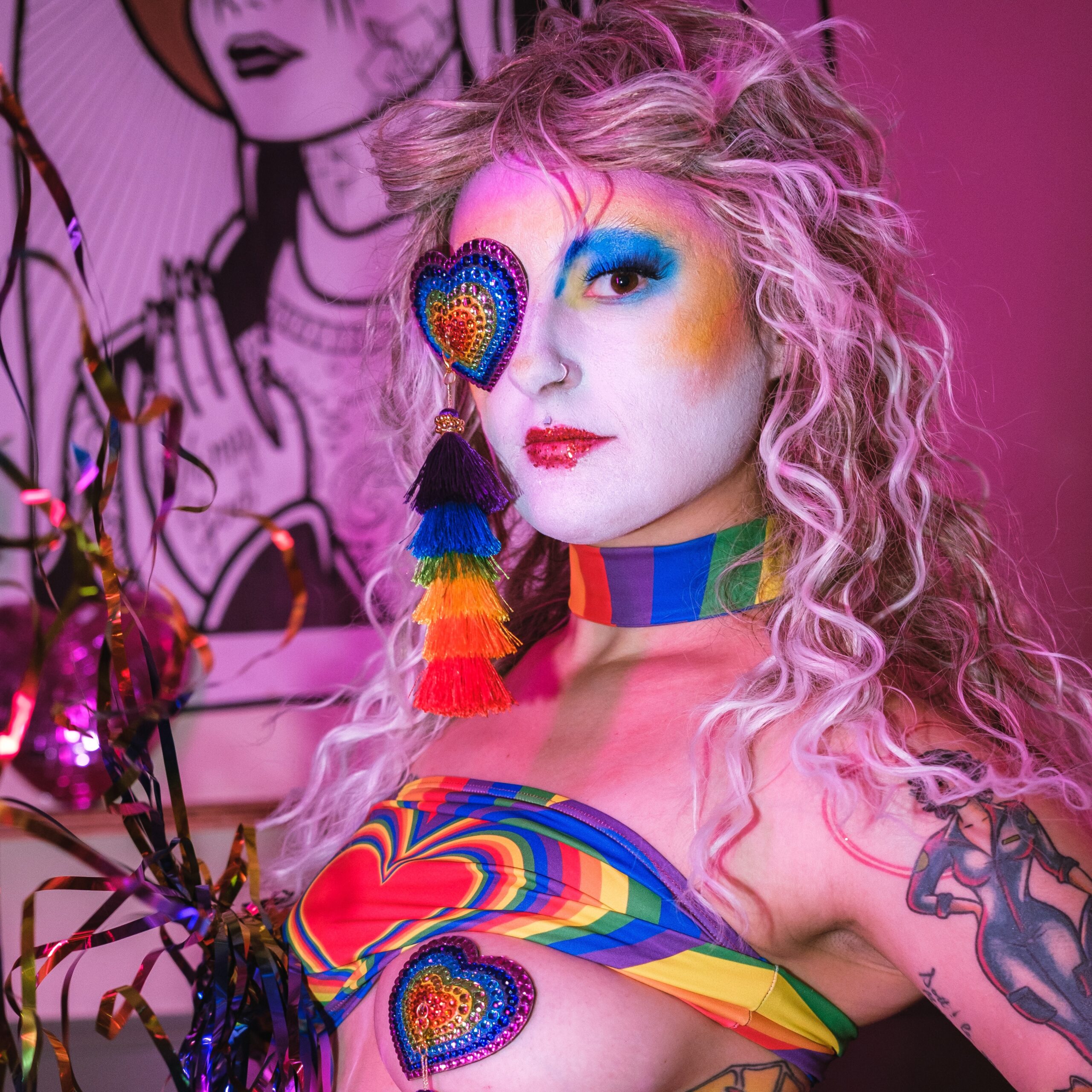Saccage Dorée en tenue burlesque arc-en-ciel, maquillage coloré et cache-œil en cœur pailleté, posant dans une lumière rose vive – Photo Léo White