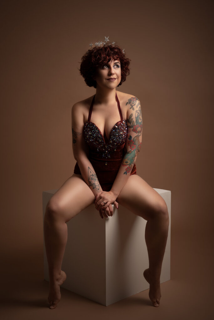 Betty Be Good assise en pied sur cube blanc, bustier rouge brodé, cheveux roux bouclés, tatouages visibles, ambiance chaleureuse – Photo Studio Bulles d'Amour