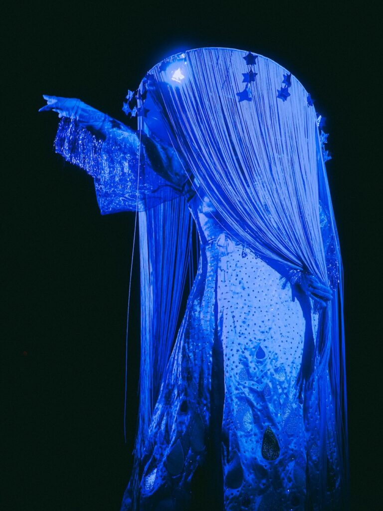 Maureen Blue Lagoon en costume burlesque bleu lumineux, entièrement recouverte de franges et de strass, bras tendu sur scène dans l’obscurité – Photo Léa et Dimitri Photographie.