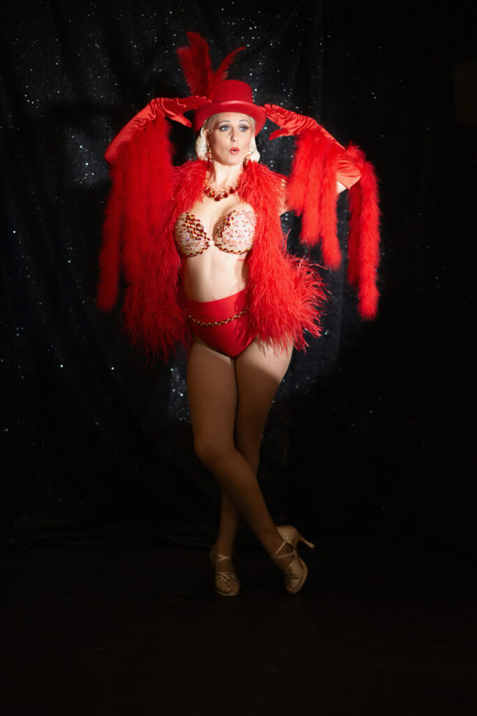 Artiste burlesque Suzy Champagne en costume rouge à plumes, portant chapeau et boa assortis sur scène devant un fond noir pailleté, photographiée par St production 79.