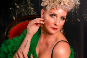 Artiste burlesque Suzy Champagne en robe de tulle verte, posée sur un fauteuil, coiffure blonde structurée et accessoire floral doré, photographiée par The Trustmaker.