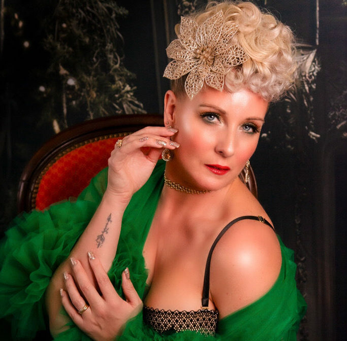 Artiste burlesque Suzy Champagne en robe de tulle verte, posée sur un fauteuil, coiffure blonde structurée et accessoire floral doré, photographiée par The Trustmaker.