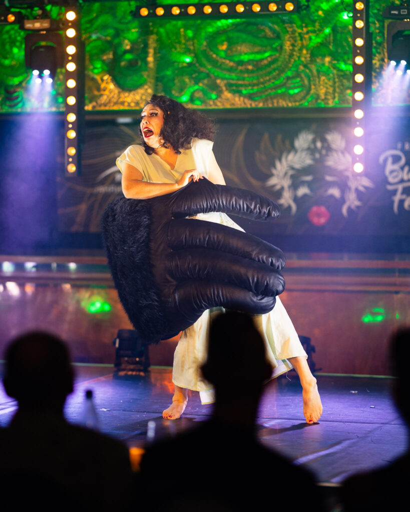 Rita Noworth sur scène, vêtue d’une robe claire fluide, manipulant une grande main noire rembourrée, devant un décor lumineux vert, vue depuis le public.