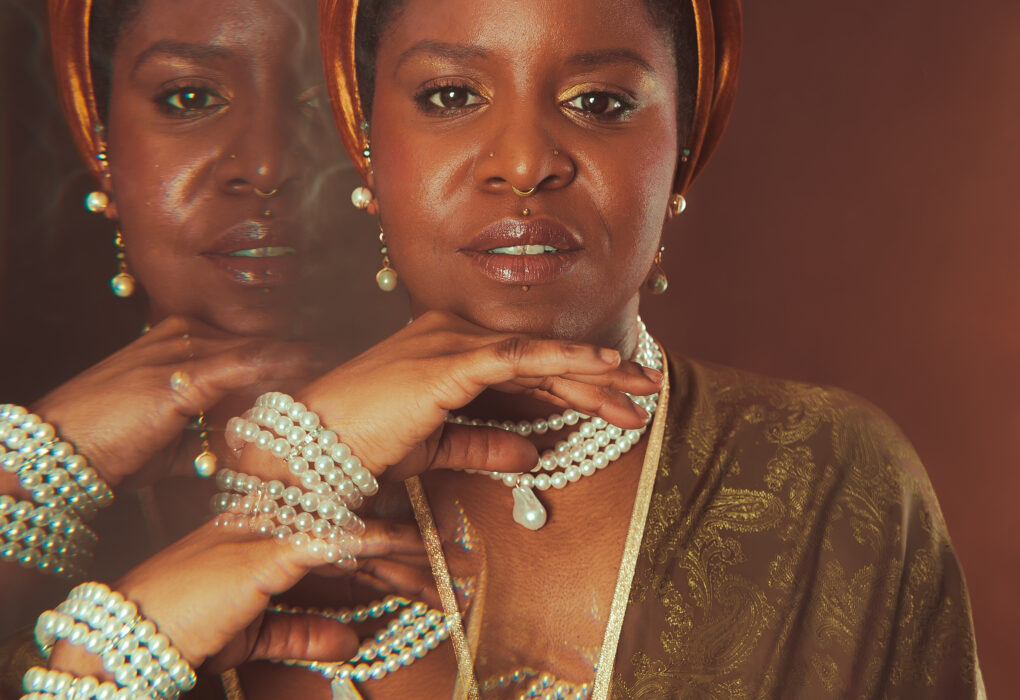 Gros plan artistique de Calie Tee portant un turban en velours, plusieurs colliers et bracelets de perles, avec effet de surimpression des mains sur fond brun.