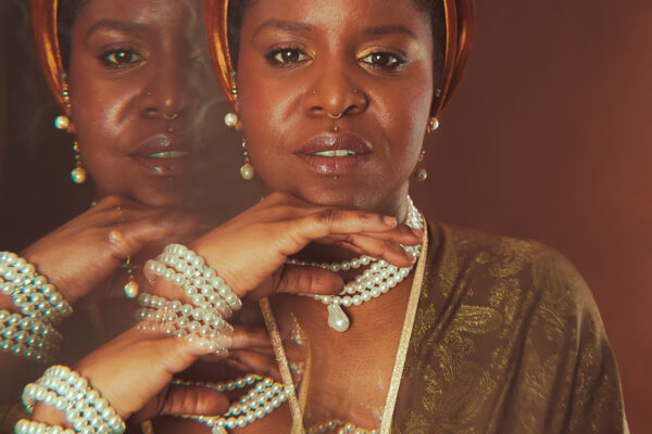 Gros plan artistique de Calie Tee portant un turban en velours, plusieurs colliers et bracelets de perles, avec effet de surimpression des mains sur fond brun.