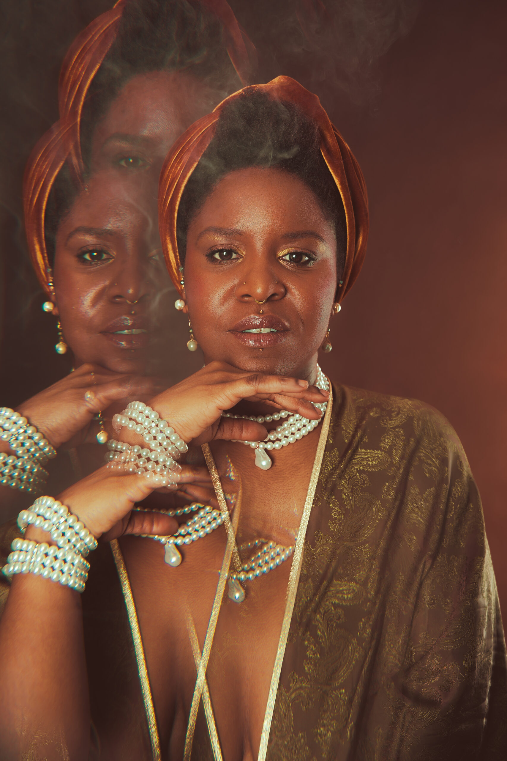 Gros plan artistique de Calie Tee portant un turban en velours, plusieurs colliers et bracelets de perles, avec effet de surimpression des mains sur fond brun.