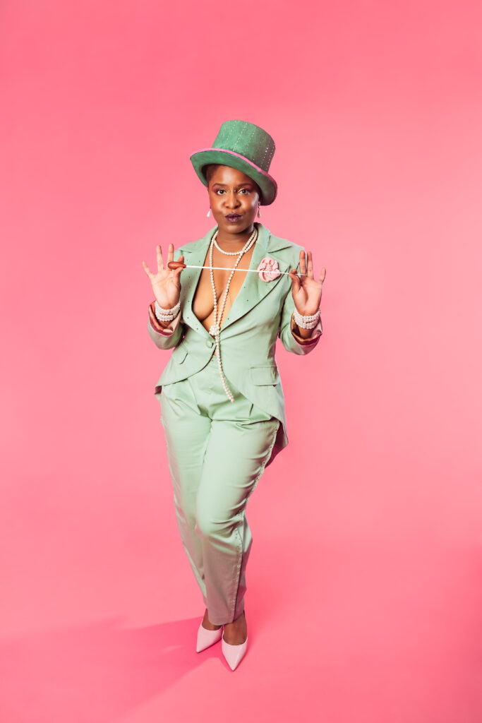 Artiste burlesque Calie Tee en costume trois-pièces vert pastel, chapeau haut-de-forme assorti et escarpins blancs, posant devant un fond rose uni.